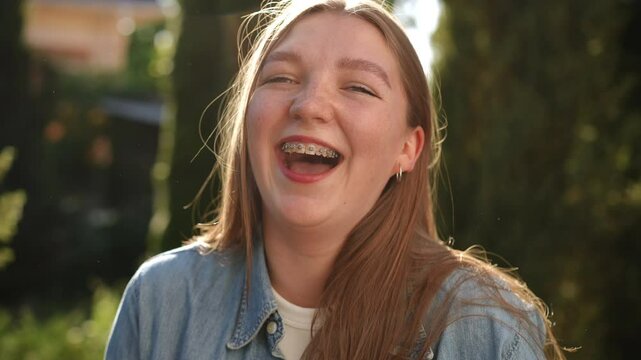 Close-up. A girl with braces and long hair looks at the camera, smiling widely and laughing hard, standing outdoors on a sunny summer day
