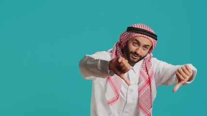 Arabic person in historic dress and kufiyah gives thumbs down and indicates discontent with what he sees. In studio, islamic adult showing evident rejection and negative signal.