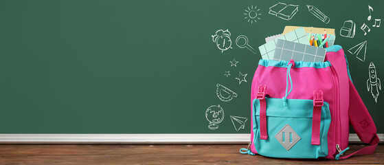 Colorful school backpack with notebooks and pencils on wooden table near green chalkboard