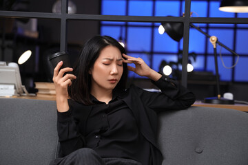 Tired Asian businesswoman with cup of coffee sitting on sofa in office