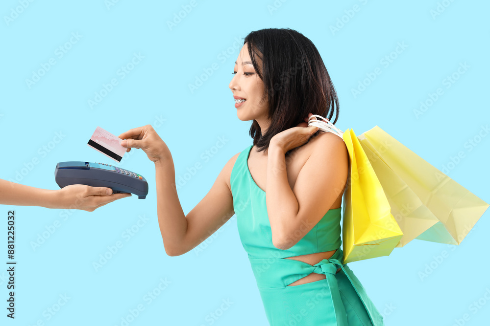 Poster happy stylish young asian woman with shopping bags and credit card paying via terminal on blue backg