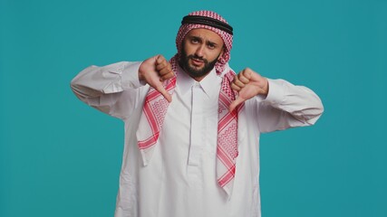 In front of background, muslim adult gives thumbs up indicating fulfillment and happiness while dressed in traditional arabic clothes. Young man wearing religious islamic clothing, like sign.