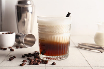 Glass of White Russian cocktail and coffee beans on white tile background