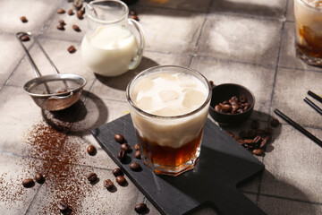 Glass of White Russian cocktail with coffee beans and jug of cream on grey tile background