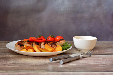 Grilled sausages with potatoes, cherry tomatoes, herbs and hot peppers on a large ceramic plate, cutlery and sauce nearby.