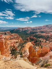 the breathtaking landscape of Bryce Canyon with its iconic red rock formations and vast, rugged terrain under a bright blue sky with scattered clouds. The panoramic view highlights the natural beauty 