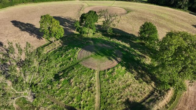 The Navan Fort, County Armagh, Northern Ireland, September 2022. Drone orbits left closely above cultural heritage site on top of circular hilltop descending below tree line to reveal ritual area.