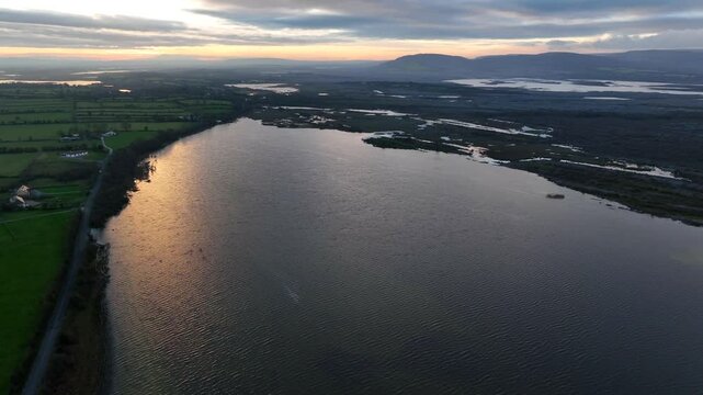Lough Bunny, County Clare, Ireland, November 2023. Drone slowly pulls back above lake water shimmering as it reflects dusk light descending to shoreline.