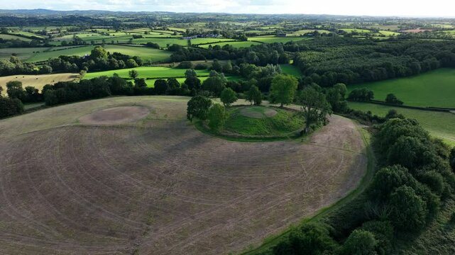 The Navan Fort, County Armagh, Northern Ireland, September 2022. Drone orbit left above circular hilltop retreating back as grassy landscape is covered by large cloud shadow.