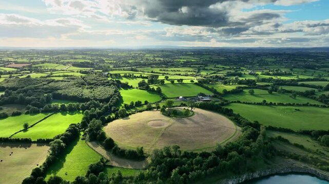 The Navan Fort, County Armagh, Northern Ireland, September 2022. Drone orbits around grassy knoll with vibrant bright green fields and lush forests on a sunny day.