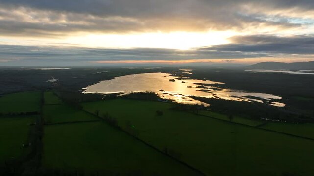 Lough Bunny, County Clare, Ireland, November 2023. Drone orbits lake in the Burren as golden sunset rays illuminate lake amongst farmland fields.