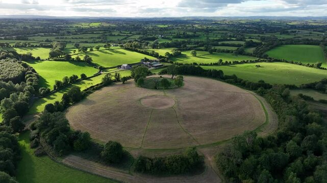 The Navan Fort, County Armagh, Northern Ireland, September 2022. Drone orbit right as cloud shadow disappears across grassy hillside with vibrant green fields around.