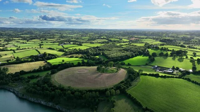 The Navan Fort, County Armagh, Northern Ireland, September 2022. Panoramic drone overview of circular grassy hilltop with large cloud shadow passing over top as sunlight breaks between clouds in sky.