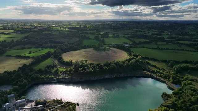 The Navan Fort, County Armagh, Northern Ireland, September 2022. Drone orbits left above sparkling water as cloud shadow casts over dramatic lush vibrant green landscape.