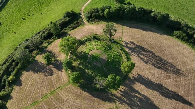 The Navan Fort, County Armagh, Northern Ireland, September 2022. Drone high angle pullback and tilt up from grassy knoll with tree shadows cast along hillside in vibrant green fields.