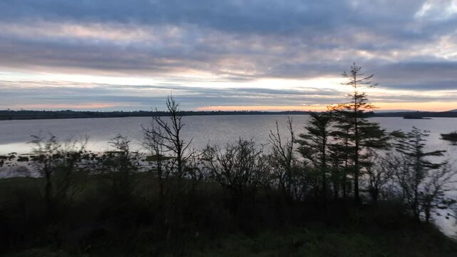 Lough Bunny, County Clare, Ireland, November 2023. Drone dolly above scraggly trees on lake shore with shimmering windswept water under dramatic blue hour sunset sky.