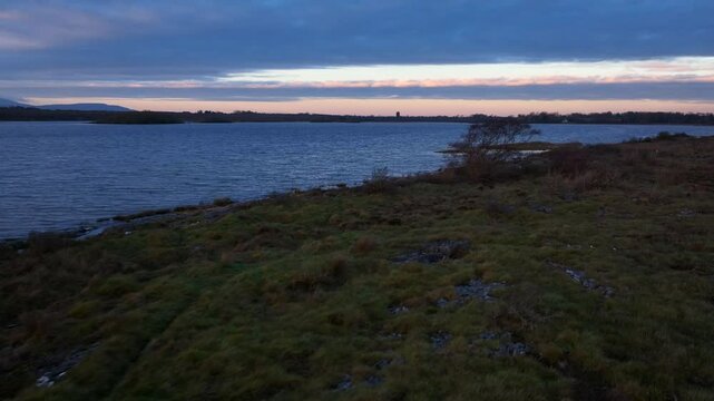 Lough Bunny, County Clare, Ireland, November 2023. Drone flies low along lakeshore edge with shrubs growing on rocky shoreline in the Burren at blue hour.