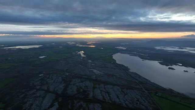 Lough Bunny, County Clare, Ireland, November 2023. Drone soars high in sky above rocky textured landscape of the Burren with last light of sunset glow on horizon.