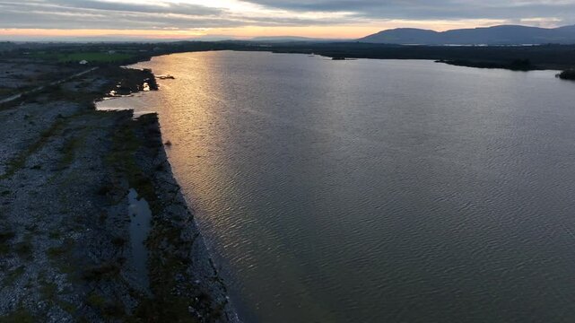 Lough Bunny, County Clare, Ireland, November 2023. Drone reverse dolly along lakeshore edge tilting up to reveal beautiful beam of sunlight light shimmering on open water.