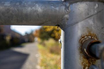 Close-up view captures rusty metal barrier with peeling paint - single water droplet poised at edge - against a backdrop of blurred autumnal trees lining a pathway. Taken in Toronto, Canada.