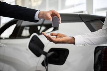 Dealer handing car key to businessman with electric SUV in background inside car dealership,...