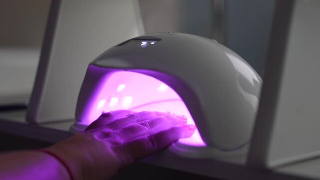 Woman puts hand with fresh beautiful pink modern gel polish manicure into led uv lamp for curing top cover of nail polish. Woman dries her nails in a professional UV lamp