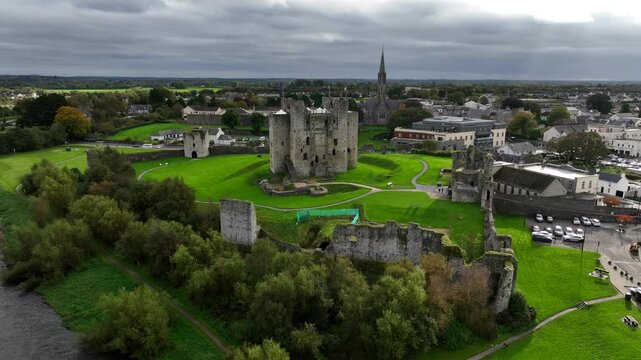 Trim Castle, County Meath, Ireland, October 2023. Drone orbits clockwise pulling away to showcase grand lawns behind steep Medieval Fortress walls built upon hill  in the center of town.