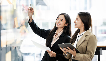 Two asian business women working together on wall glass with post it stickers. Modern startup office
