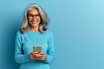 Fototapeta premium Joyful Senior Woman with Glasses Using Smartphone on Blue Background. Horizontal banner with copy space