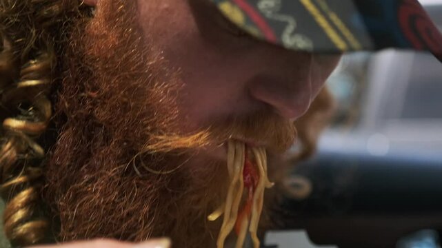 Red bearded guy eats Chinese udon with meat and vegetables in wok close up with chopsticks, chews takeaway food with appetite.