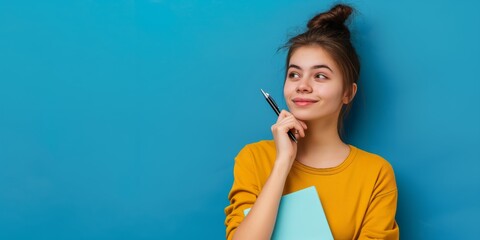 Fototapeta premium A young woman is deep in thought while holding a notebook against a blue background. She appears focused and inspired, possibly brainstorming or planning for her academic pursuits