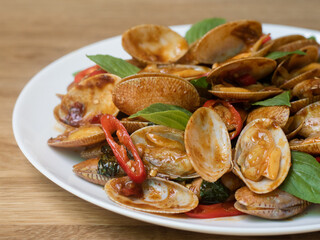 Stir fried striped clams with basil, chilli paste, garlic and fresh chilies. Placed in a white plate with a beautiful pattern and on the wood pattern background.