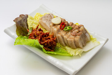 Close-up of boiled pork with napa cabbage, lettuce, seasoning sauce, red and green peppers on square dish and white floor, South Korea
