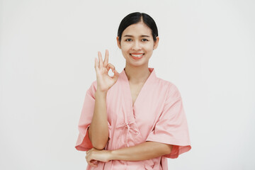 A young Asian female wearing a pink patient uniform stands isolated against a white wall...