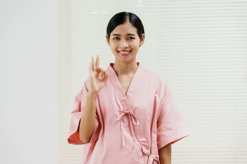 A young Asian female wearing a pink patient uniform stands isolated against a white wall...