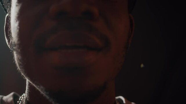 Closeup of confident young Black man with goatee demonstrating heavy silver rapper chain on his neck at camera while standing on dark studio background