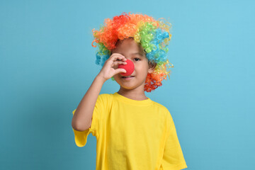 Cute little asian boy in clown wig, hand holding clown nose isolated on blue background. copy space