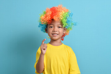Cute little asian boy in clown wig isolated on blue background. index finger up. copy space