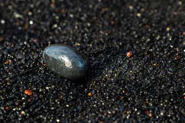 A small and wet black rock is laying on the ground of a black sanded beach