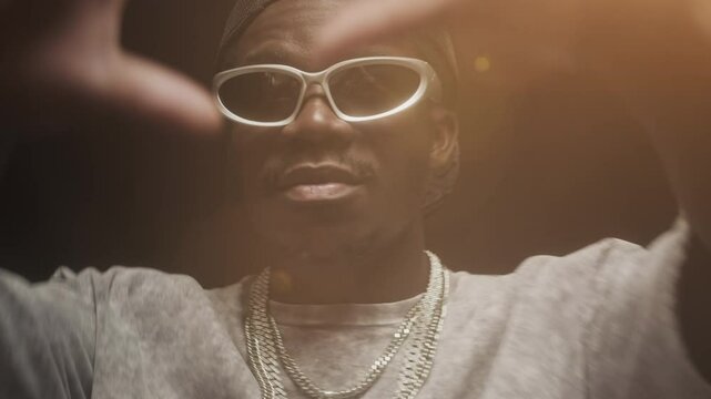 Chest up of cool African American male rapper wearing stylish glasses, backwards cap and chain necklace dancing to music at party on black background with orange source of light behind him