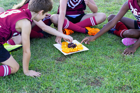 children in sports uniform sitting on ground and having fruit off a plate on the grass