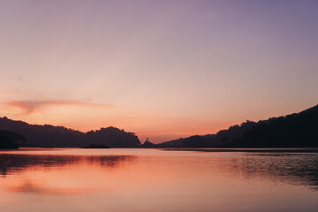 Fototapeta premium A beautiful sunset over Sermo lake, Yogyakarta, with a small island in the middle. The sky is a mix of pink and purple hues, creating a serene and peaceful atmosphere.
