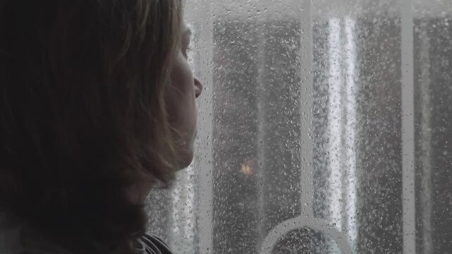 A close-up side view of a woman with dark hair, leaning sadly on a window, reflecting her sorrow as the storm rages outside. The scene captures a poignant moment of sadness and reflection.