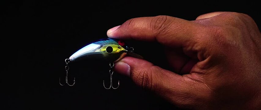 Hand holding artificial lure of mini minnow with treble hook installed on black background.