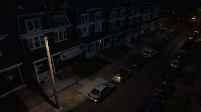 Aerial night view of a quiet residential street with row houses and parked cars. A single streetlight illuminates part of the sidewalk and road, creating a serene and dimly lit scene.