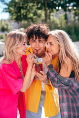 Young three stylish friends posing in the street. Fashion man and two cute female in casual summer clothes. Smiling cheerful women and guy outdoors, Hold and drink lemonade from one plastic cup