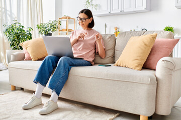 A woman in cozy homewear sits on a couch, absorbed in her laptop.