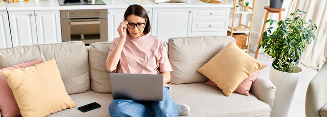 A mature woman in cozy homewear sits on a couch, engrossed in using a laptop.