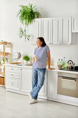 A mature woman in cozy homewear standing in a kitchen, next to a sink.