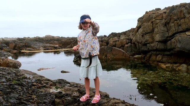 Girl with bamboo fishing pole holds up klipfish next to rockpool on coastline